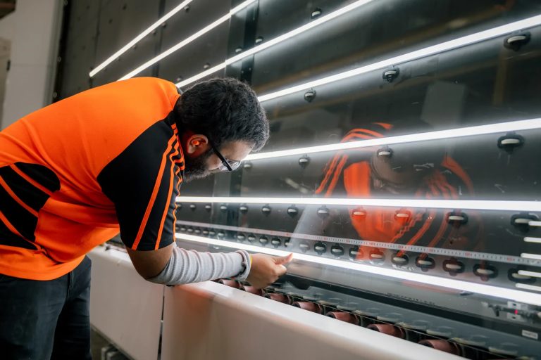 Tough Glass technician closely inspecting a glass panel at an illuminated quality-control station.
